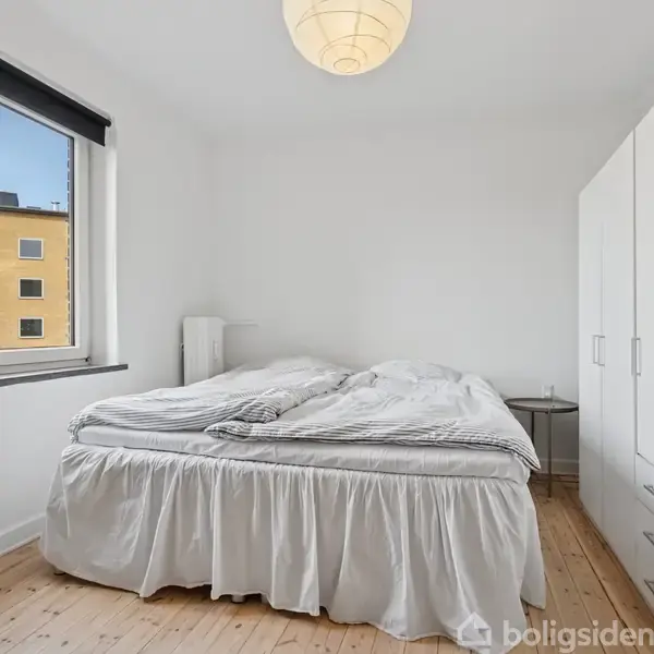 En seng med hvidt sengetøj står i midten af et lyst soveværelse. Der er et stort vindue til venstre og en hvid garderobe til højre.