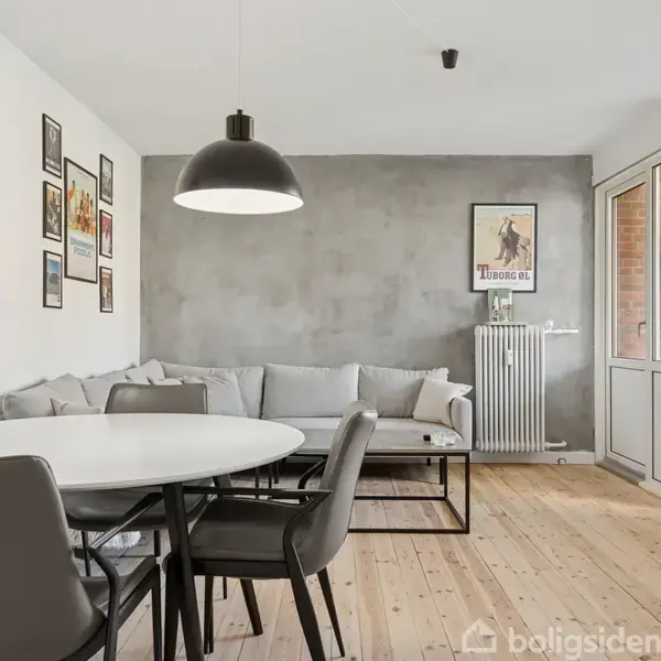 En spisegruppe med rundt bord og fire stole står i et lyst rum. En grå sofa langs væggen. Vægpynt og plakat hænger over radiator. Terrasse udsyn udenfor.