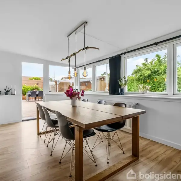 Et rektangulært spisebord med sorte stole omringes af store vinduer med udsigt til en terrasse og have. Bordet er dekoreret med en vase med blomster.