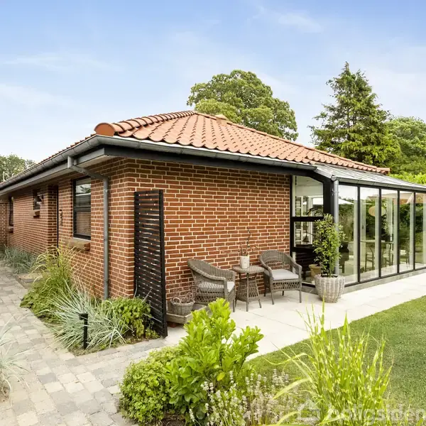 Rødt murstenshus med rødt tegltag, har en glasveranda og udendørs siddepladser. Huset står i en velplejet have med flisebelagte stier og forskellige planter, omgivet af grønne træer og buske.