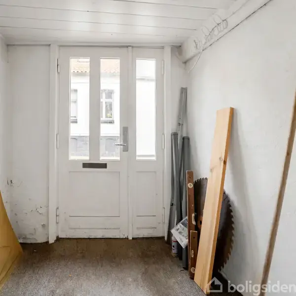 Dørparti med glasruder står uåbnet i et lille, hvidmalet rum. Væggene er omkranset af plader og værktøj, som en savklinge og træplanker, der antyder opbevaring eller renovering.