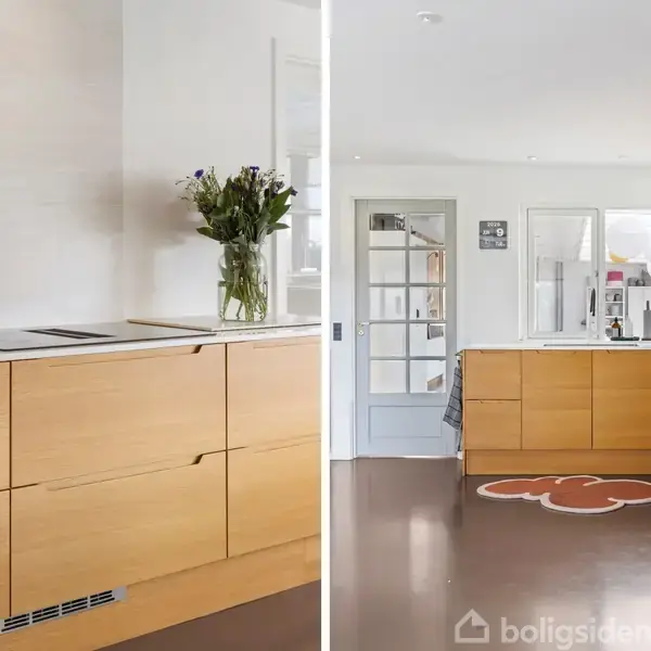 Moderne køkken med lyse træskabe og hvide vægge. Blomster står i en vase på bordet, og der er åbne hylder til opbevaring. Lysindfald gennem store vinduer.