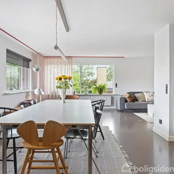 Spisebord med stole står i et lyst køkken-alrum. En vase med solsikker er i midten af bordet. Vinduer på venstre side giver naturligt lys. Sofa i baggrunden.