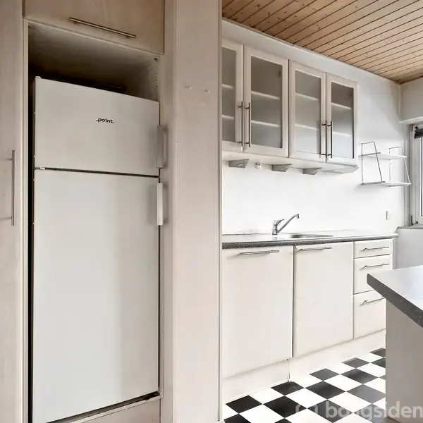 Et hvidt køleskab står ved siden af køkkenskabe, mens et sort-hvidt skakternet gulv strækker sig under dem i et lyst køkken med træloft og vindue.