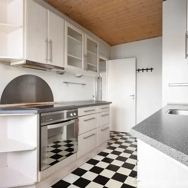 Et moderne køkken med lyse skabe og en indbygget ovn står i et rum med sort-hvid ternet gulv og træloft. En vask er placeret på en mørk bordplade.