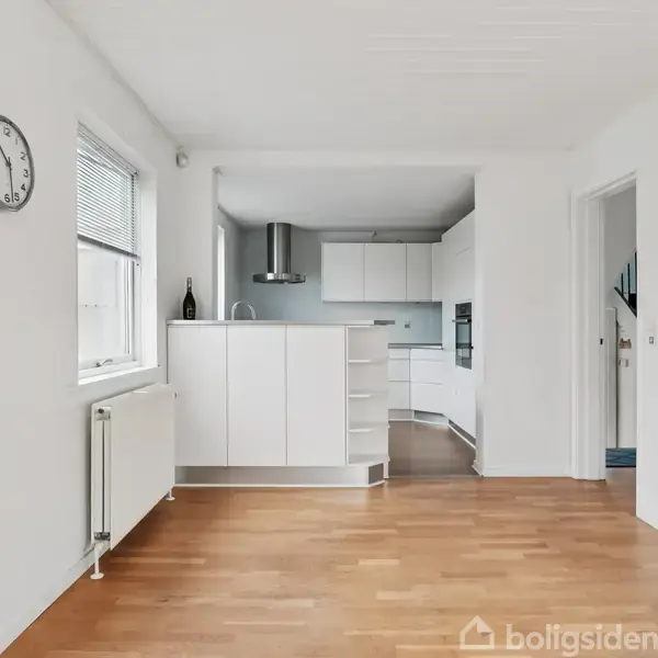 Køkken med hvide skabe og stålemhætte, ingen personer til stede. Trægulv fører ind til køkken, hvidmalede vægge. Vægur til venstre, vindue og radiator ved siden af.