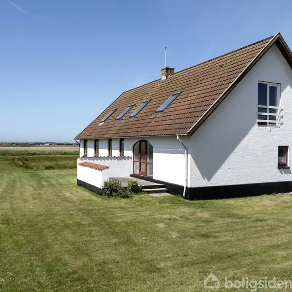 Et hvidt hus med rødt tag står på en grøn græsplæne i et landligt område. Huset har flere vinduer, og landskabet er fladt med en klar blå himmel.