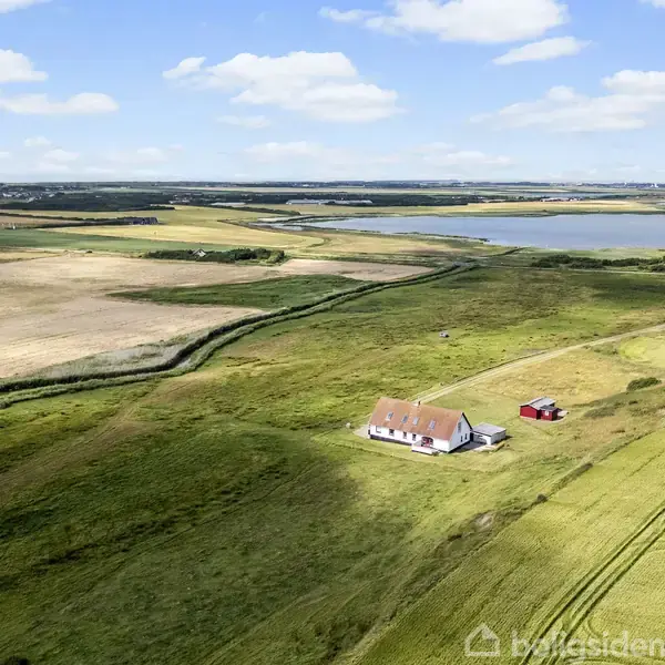 Hus står stille midt i åbent, grønt landskab med marker omkring, under blå himmel med hvide skyer. En lille, rød bygning ses ved siden af huset.