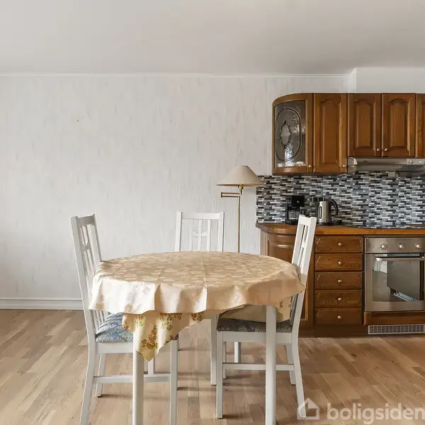 Et rundt spisebord med gul dug står i en stue-køkkenkombination. Køkkenet har brune skabe og mosaik-fliser bagved, komplet med ovn og mikrobølgeovn. Fire hvide stole omkranser bordet.