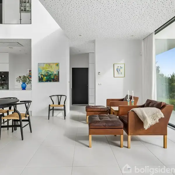 Stue med moderne møbler. Sofa og stole er arrangeret ved siden af store vinduer. Bord med stole til venstre, kunst på væggene. Åbent og lyst rum med udsigt til natur.