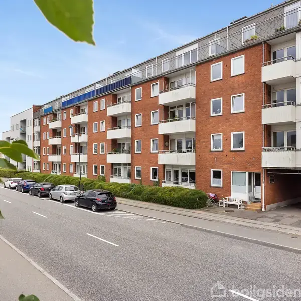 En række etagebygninger med røde murstensfacader og balkoner står langs en stille vej. Forskellige biler er parkeret langs fortovet, og grønne blade ses øverst i billedet.