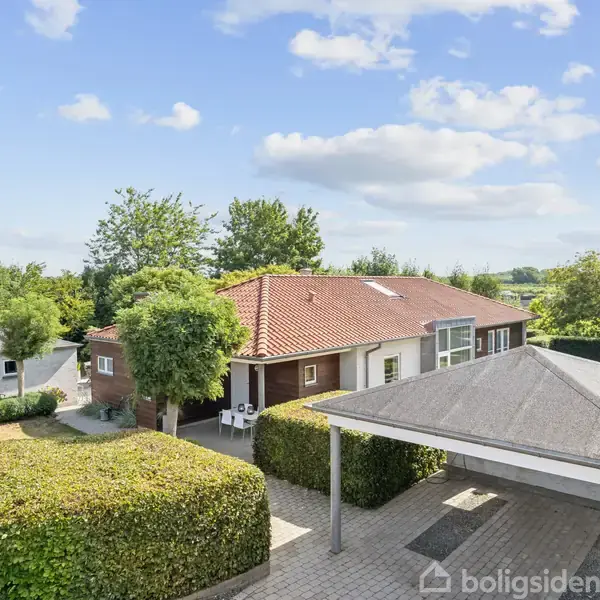 Hus med rødt tegltag og udestue omkranset af grønne hække og træer i en have. Foran huset ses en carport. Himlen er blå med skyer.