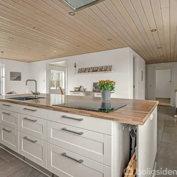 Et moderne køkken med en stor central køkkenø med vask og kogeplade. Lys træbeklædning i loftet og stenvægge skaber en lys og åben atmosfære. Flere vinduer slipper dagslys ind.