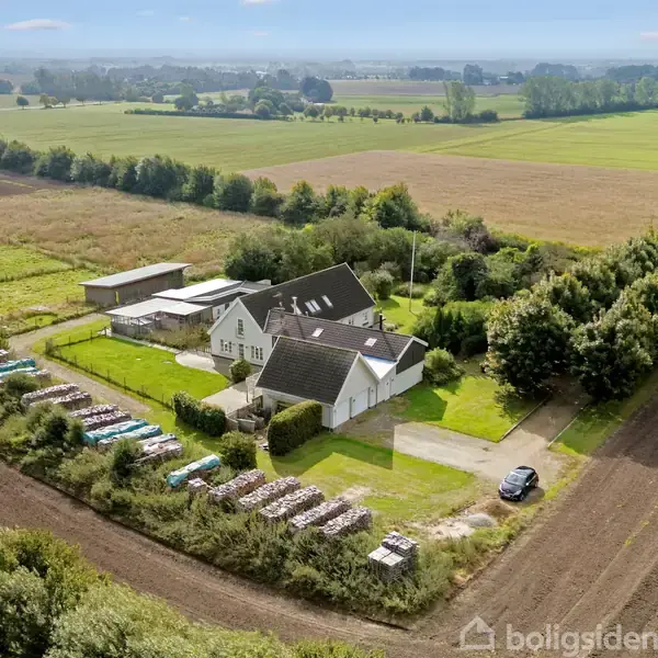 Hus med mørkt tag omgivet af grønne træer og marker. Flere opbevaringsområder med brændestabler og landbrugsudstyr. Markveje og en parkeret bil i landligt landskab.