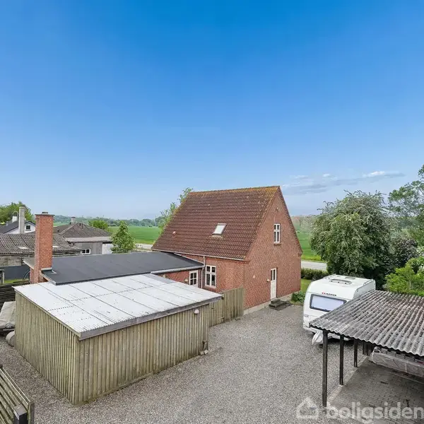Et rødt murstenshus med skråt tag står på en grusbelagt grund. Rundt omkring huset ses en carport, en campingvogn og et skur. Baghaven har grønne træer og marker.