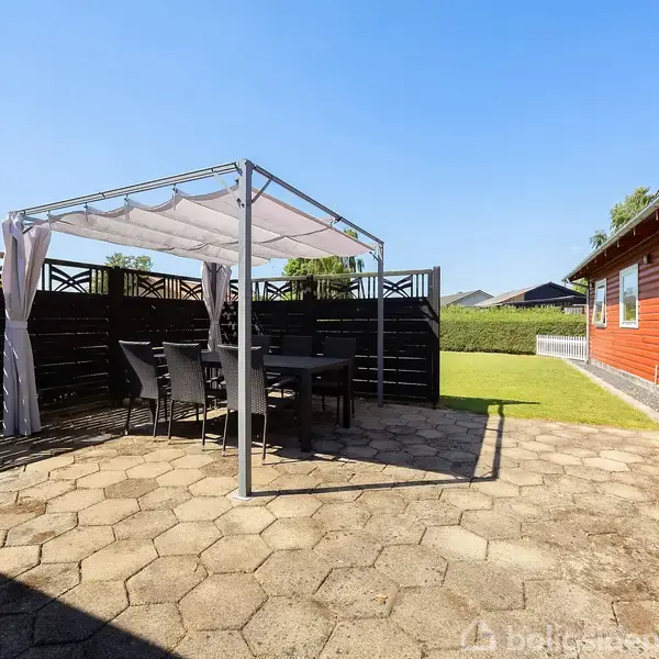 Pavillon med hvidt solsejl står på flisebelagt terrasse. Sorte stole og bord er placeret under sejl. Til højre ses en rød bygning med vinduer. Græsplæne i baggrunden.