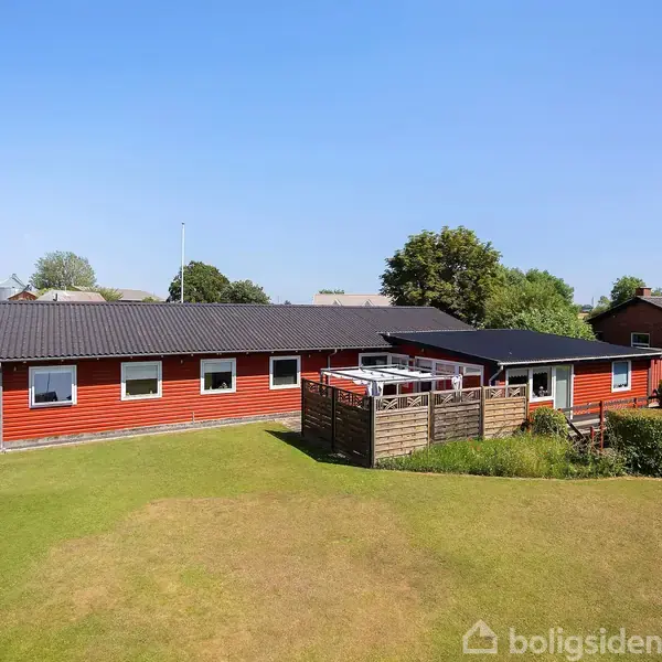 Rødt, en-etages hus står stille i en grøn have med få træer. En terrasse med hæk omgiver huset, og himlen er klar og blå.