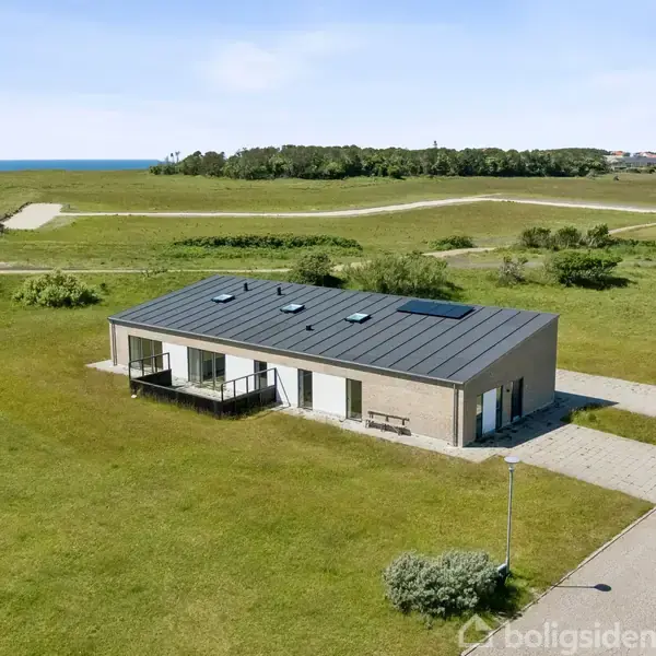 En moderne, enkel bygning står stille på en velholdt græsplæne. I baggrunden er der udsigt over et åbent landskab. Der er flisebelægning foran bygningen og en asfalteret sti til indgangen.