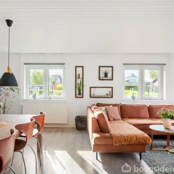Sofa i hjørneform med brændte orange puder står i en stue. Foran sofaen står et rundt sofabord. Omkring er hvide vægge, vinduer og spisestuemøbler i træ.