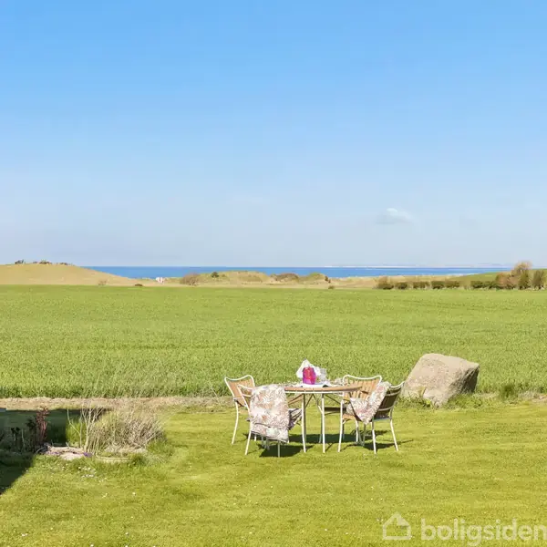 Et bord med stole står på en grøn græsplæne. Bagved ses en stor mark og horisonten med blå himmel og hav i baggrunden.