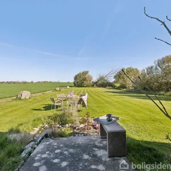 Terrasse med et bord og stole står på en grøn græsplæne. Omkring er der åbne marker og træer under en klar blå himmel. Ingen tekst er synlig i billedet.