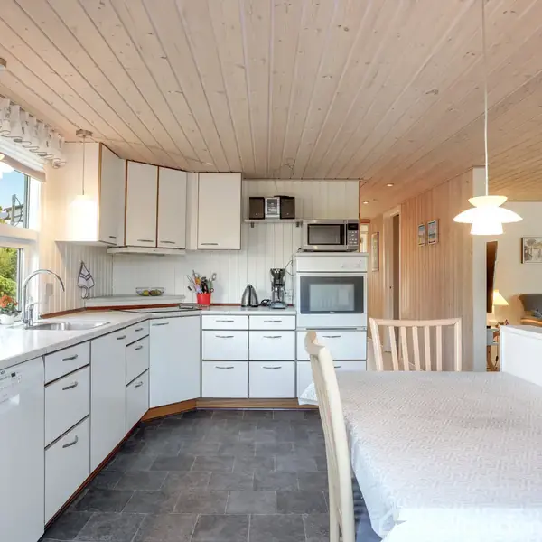 Et lyst køkken med hvide skabe og en indbygget ovn. Et spisebord med stole står til venstre. Store vinduer giver udsigt til grønne træer udenfor.