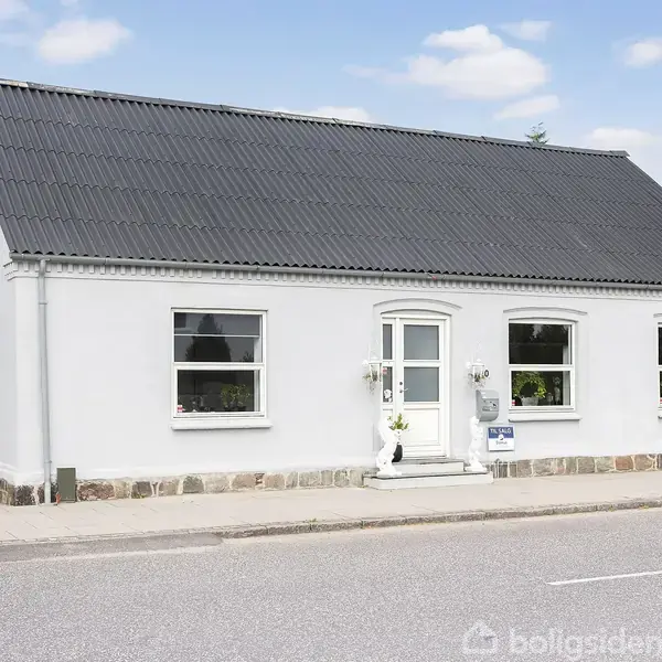 En hvid bygning står stille langs en vej, med tre vinduer og en central indgangsdør. Den har et sort tag og små dekorationer foran. En bil ses parkeret til venstre.