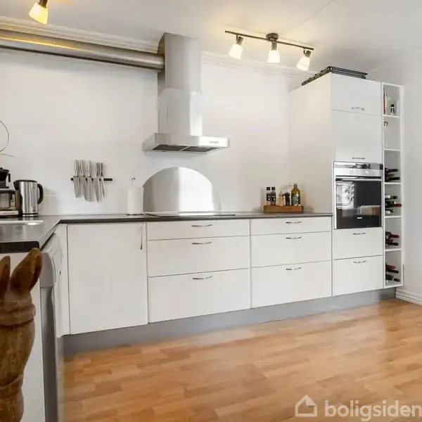 Hvidt køkken med skuffer og skabe, en emhætte over komfuret, placeret i et moderne rum med trægulv. En træhest er i forgrunden, planter og redskaber på bordet.