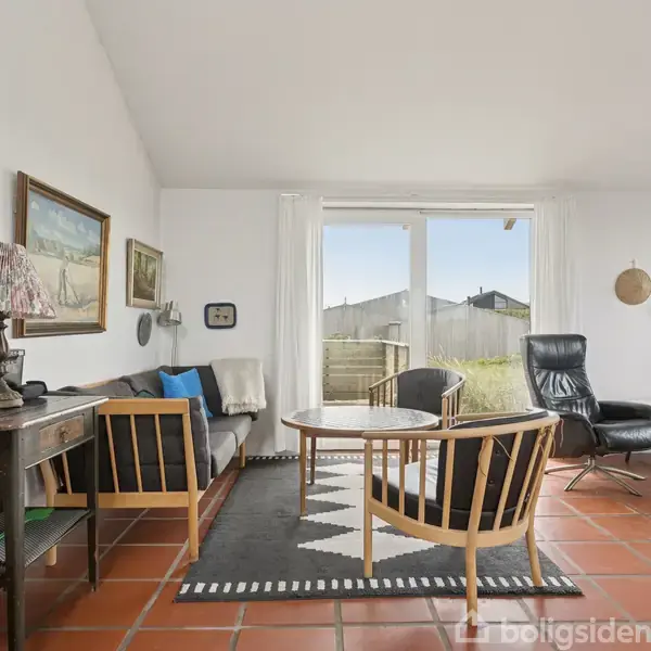 Stue med træmøbler arrangeret omkring et rundt bord på en sort-hvid stribet tæppe. Store vinduer viser udsigt til en terrasse og have. Indretningen er lys og minimalistisk.