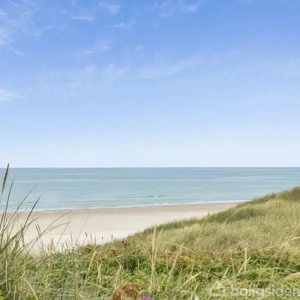 Strand set mod blå himmel, med bølger, blide klitter og marehalm i forgrunden skaber en fredelig kystoplevelse.