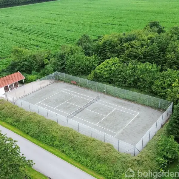 Tennisbane omgivet af hegn står stille ved siden af en lille hvide bygning med rødt tag, omringet af grønne marker og træer, ved vejkant i landligt område.
