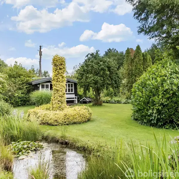 Et lille, sort-hvidt hus står i baggrunden, omgivet af en frodig have med grønne buske og træer. En dekorativ dam med siv og åkander ligger i forgrunden.