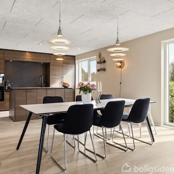 Et spisebord med sorte stole står i en moderne spisestue. Et køkken med træpaneler og sort udstyr ses i baggrunden. Store vinduer tillader udsigt til haven.