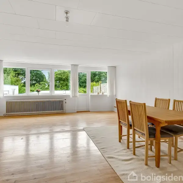 Et spisebord med stole står på et tæppe i en lys og rummelig stue med trægulv. Store vinduer åbner mod en grøn, frodig udsigt udenfor.