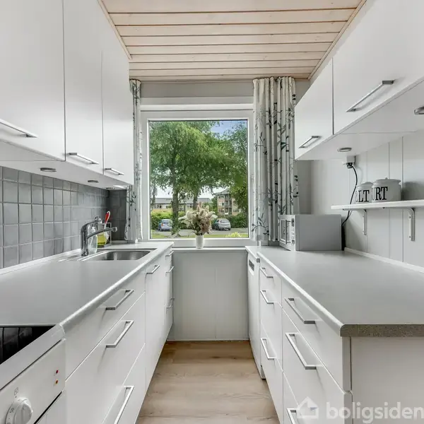 Et lyst, moderne køkken med hvide skabe og grå fliser bag komfuret. En stor vindue giver udsigt til træer udenfor. Kogeplader, vask og køkkenredskaber er synlige.