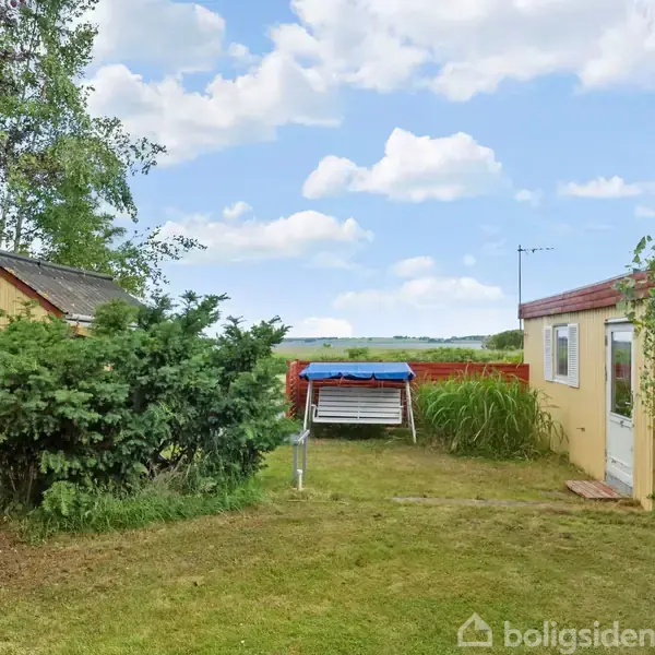 To små bygninger står i en have med græs, omgivet af buske og træer. I midten ses en blå vippe foran en udsigt over åbne marker og himmel.