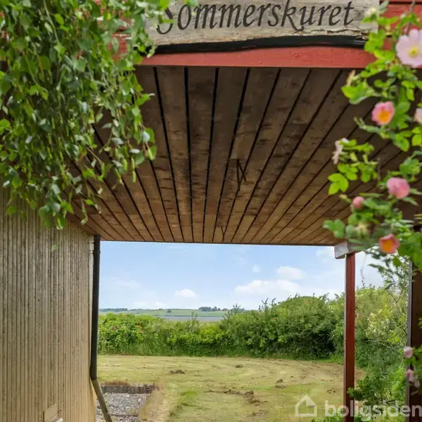 Et træskilt med teksten "Sommerskuret" hænger over en træterrasse. Foran er en grøn græsplæne omgivet af buske og blomstrende planter, med udsigt til en landlig horisont.