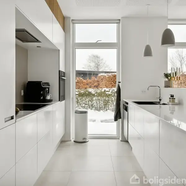 Et moderne køkken med hvide skabe og en sort bordplade, står tomt og oplyst af naturligt lys fra store vinduer. Udenfor ses en hæk og sneklædt landskab.