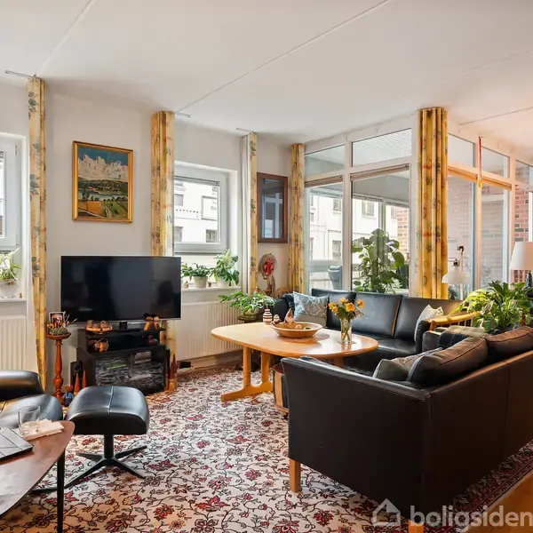 Stue med sort lædersofa og lænestol, tv på skænk, og træbord med blomster. Store vinduer med gulv-til-loft-gardiner og tæppe i blomstermønster. Indendørs planter ved vinduet.