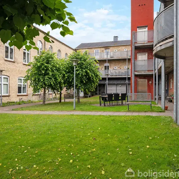 En grøn gårdhave med græsplæne og flere træer. Omkring gårdhaven ses bygninger med murstensfacader og altaner. En gruppe sorte stole og et bord står midt i græsset.