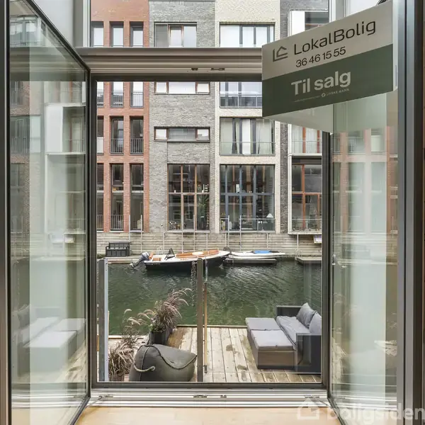 En smal altan med sofa vender ud mod en kanal med både. Gennem døre ses moderne bygninger. Skilt fra "LokalBolig" viser telefonnummer 36 46 15 55 og teksten "Til salg".