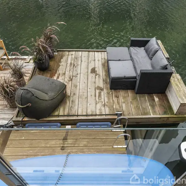 En flydende terrasse med sofa og parasol i en kanal. Træstole og bord ved siden af. Omgivet af grønne vandplanter og stilrent design, skaber en afslappende oase.