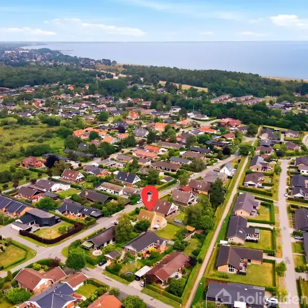 Et parcelhuskvarter set fra luften med tætliggende huse og haver, omgivet af grønne områder. En rød markering fremhæver et specifikt hus. I baggrunden ses kystlinjen og havet.