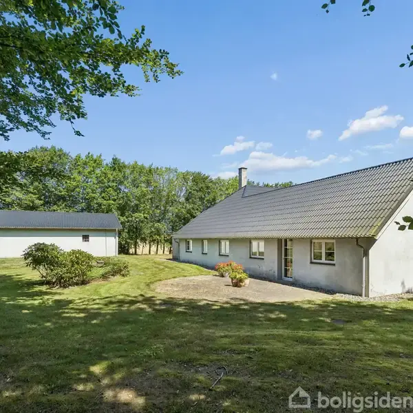 Et hvidt hus med gråt tag står stille i en grøn have omgivet af træer under en blå himmel med hvide skyer.