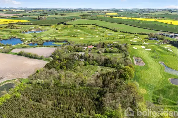 Landskab med marker og buske, omgivet af store, grønne græsarealer. Flere små søer i baggrunden og bygninger kan anes mellem træerne. Landskabet strækker sig mod en horisont af let kuperede bakker.