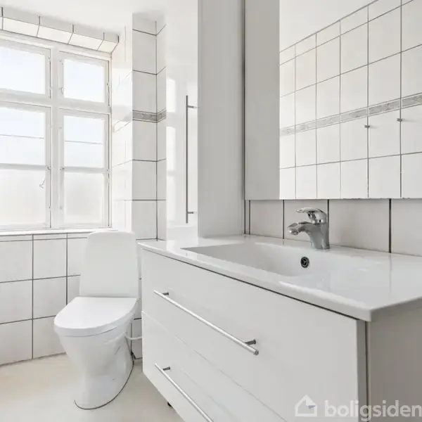 Et hvidt badeværelse med fliser, inkluderer en vask med skuffer under og et toilet ved siden af, oplyst af naturligt lys fra et vindue over toilettet.