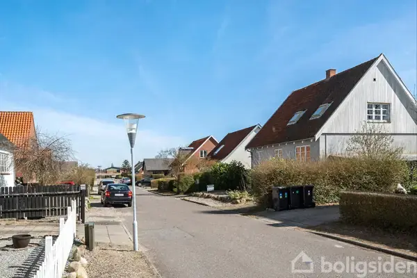 Gade med lamper og parkerede biler i et boligområde; huse med skrå tage flankerer vejen, omgivet af hække og træer under en klar blå himmel.