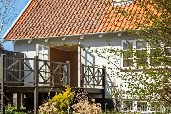 Et hus med hvid facade og rødt tegltag har en udendørs terrasse med trægulv og et stakit. Omkring terrassen er der grønne buske og planter, der skaber en naturlig ramme.