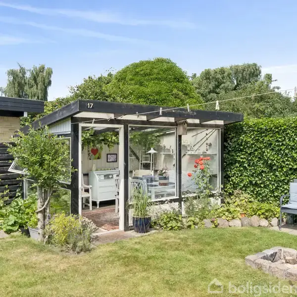 Et lille drivhus med glasvægge har planter og dekorative genstande indenfor. Det står i en grøn have med en bålplads foran. Tallene "17" er på drivhusets tag.