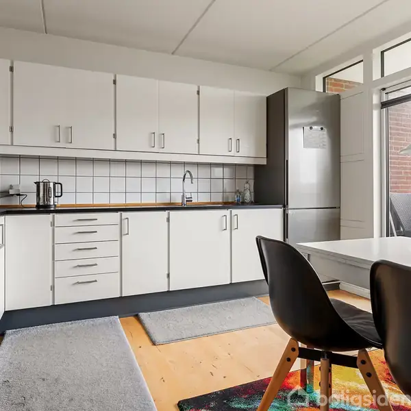 Køkken med hvide skabe og sorte greb. En stålkomfur ved siden af en grå køleskab. Køkkenbord med to sorte stole. Vinduet giver udsigt til en altan.
